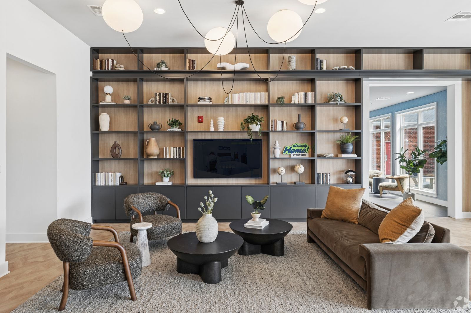 Resident lounge with a sofa, armchairs, TV, display shelving, and floor-to-ceiling windows